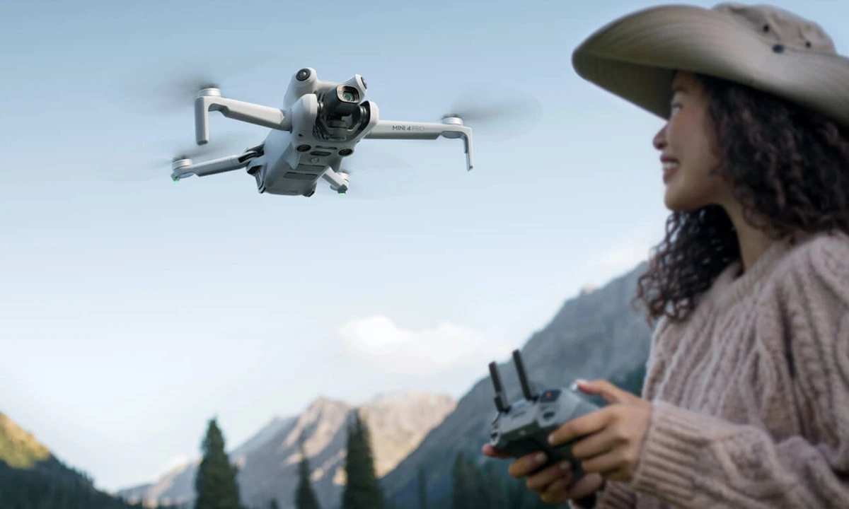 en primer plano el DJI Mini 4 Pro volando por un bosque y un poco desenfocadla una mujer con el control del dron