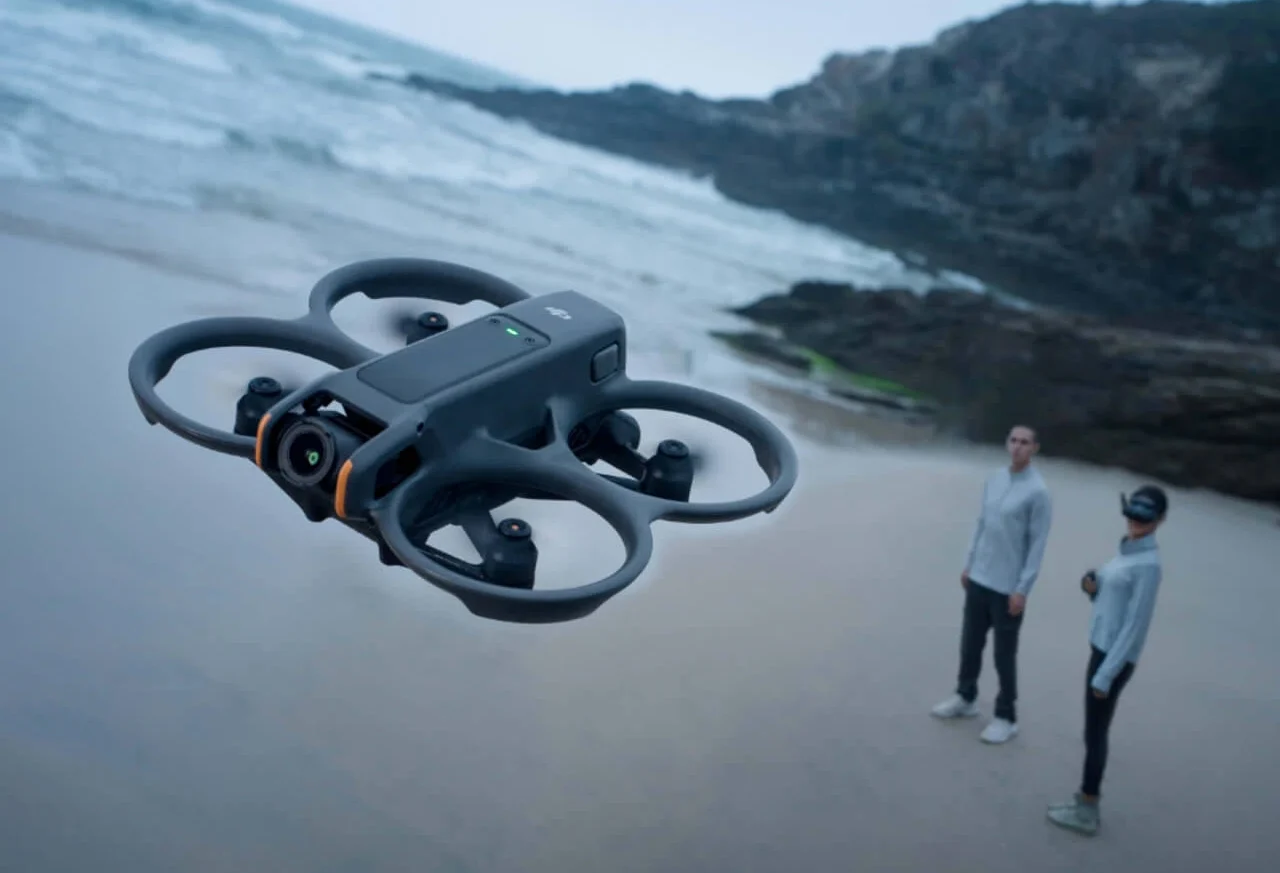 Primer plano del DJI Avata 2 volando en una playa y de fondo una persona piloteando